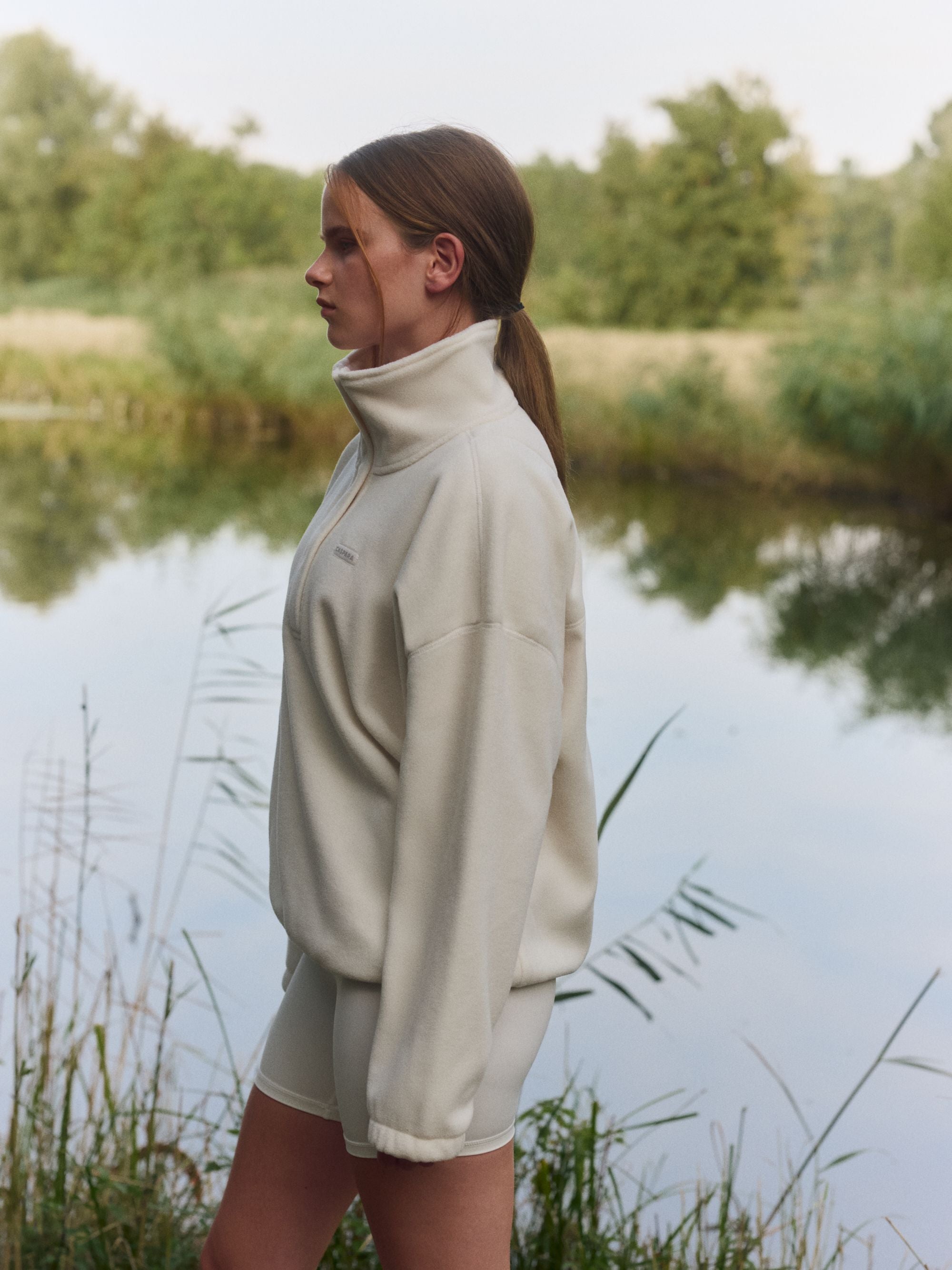 Woman wearing a creme CASPARA polar fleece half-snap pullover standing by a lake.